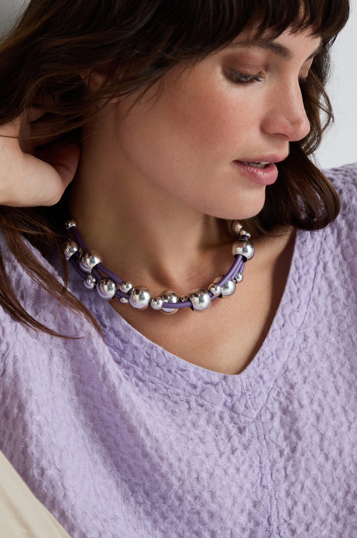 Woman in a textured lavender top wears the Infinite Spheres Necklace, which features silver acrylic spheres on violet PVC cords, as she looks down with a relaxed expression.