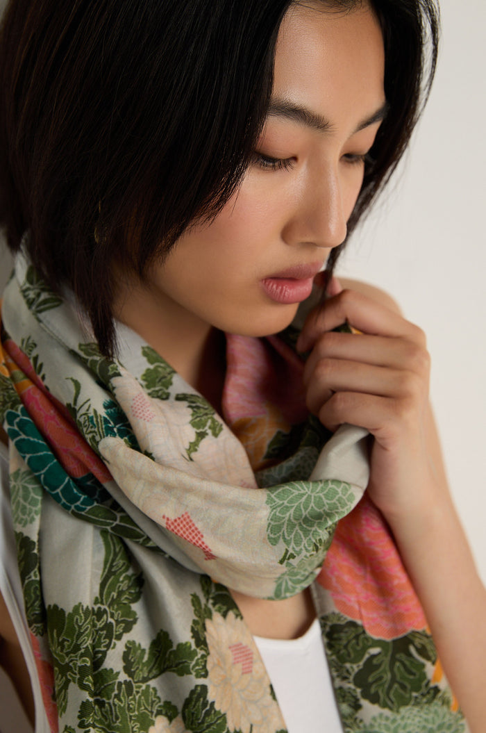 A woman with short dark hair wears the Jacquard Scarf and a white top, looking down as she touches the scarf near her face.
