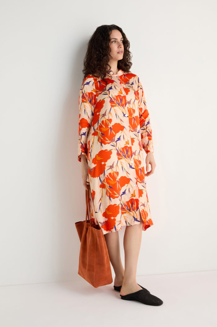 A woman stands against a white wall wearing the Japanese Floral Shirt Dress, featuring a knee-length, long-sleeve floral print with red, orange, and purple accents. She holds an orange tote bag and wears black flats.