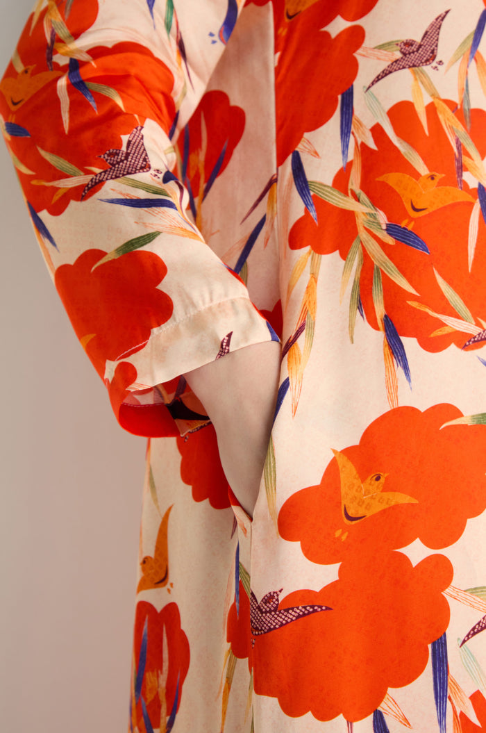 Someone wearing the Japanese Floral Shirt Dress, featuring bold orange floral and bird patterns on a light background, has their hand tucked into a side pocket.