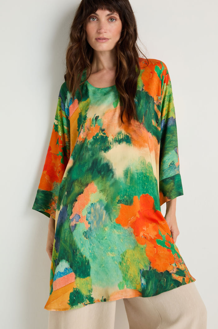 A woman stands against a plain white wall wearing the Japanese Garden Boxy Tunic, an oversized woven viscose top with an abstract green, orange, and blue pattern, paired with light beige pants.