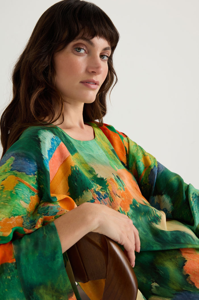 A woman with wavy brown hair wears the Japanese Garden Boxy Tunic, featuring a colorful abstract pattern, as she sits on a wooden chair and looks at the camera against a plain background.