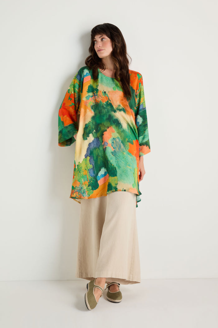 A woman stands against a white wall wearing the Japanese Garden Boxy Tunic in colorful green and orange patterns, paired with wide beige pants and olive shoes.