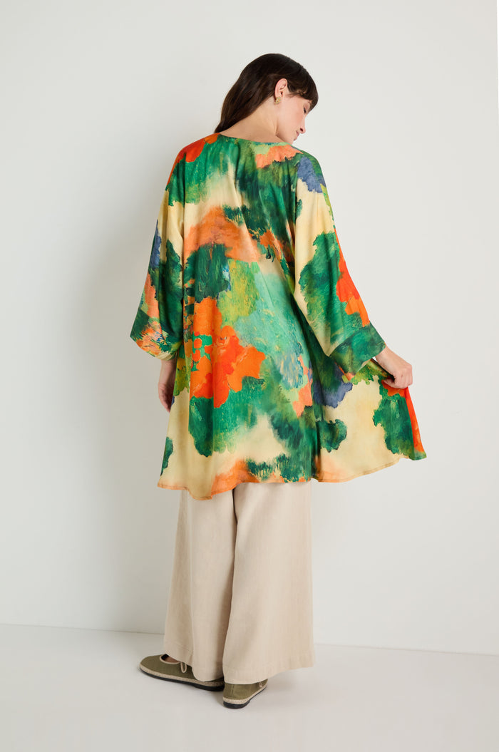 A woman stands against a white wall wearing the Japanese Garden Boxy Tunic with abstract green, orange, and yellow patterns, paired with beige wide-leg pants and green shoes.