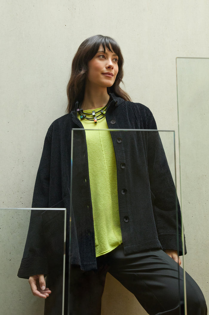 Woman standing indoors against a light-colored wall, wearing the Jumbo Cord Jacket over a lime green top and black pants, paired with a multicolored necklace.