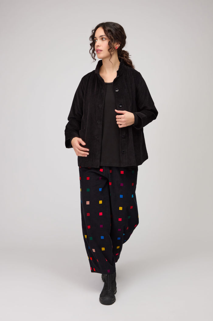 A person wearing a Jumbo Cord Jacket, black top, and loose black pants with multicolored square patterns, walking against a plain light background.