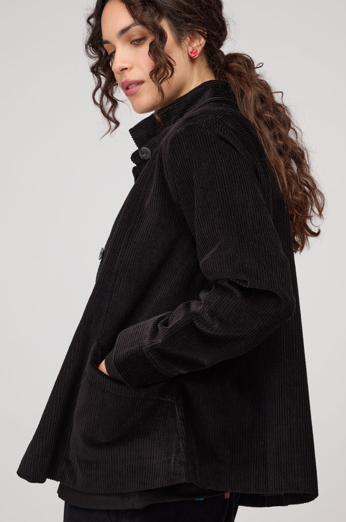 A woman with curly brown hair poses in profile against a plain background, wearing the black Jumbo Cord Jacket with one hand in her pocket.