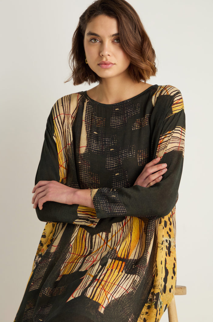 A woman with short brown hair sits with arms crossed, wearing a long-sleeved Klimt Print Easy Dress in painterly yellow, black, and red on a soft cupro-viscose blend.