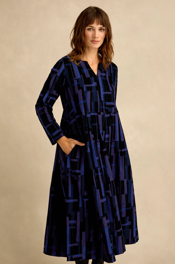 A woman wears the Layered Geometric Cord Flared Dress, a long-sleeved navy and black design with pockets, standing against a plain beige background.
