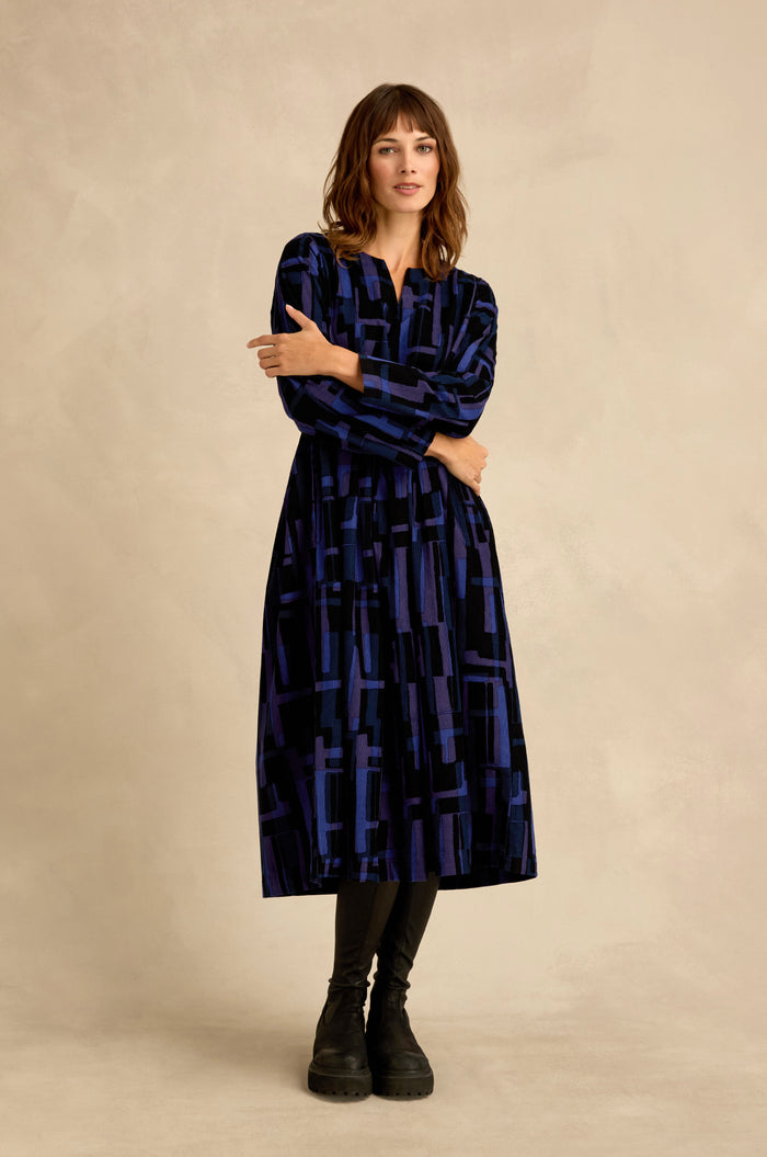 A woman stands against a neutral background, wearing the Layered Geometric Cord Flared Dress— a long-sleeve, knee-length blue and black piece featuring geometric patterns—paired with black leggings and black boots.