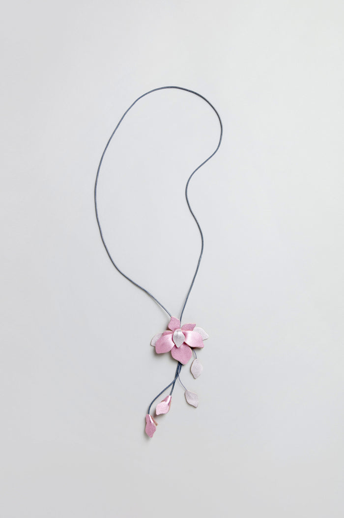 The Leather Flower Pendant is a handcrafted necklace with a long blue cord, showcasing a large pink leather flower and small pink leaf charms, all set against a light background.