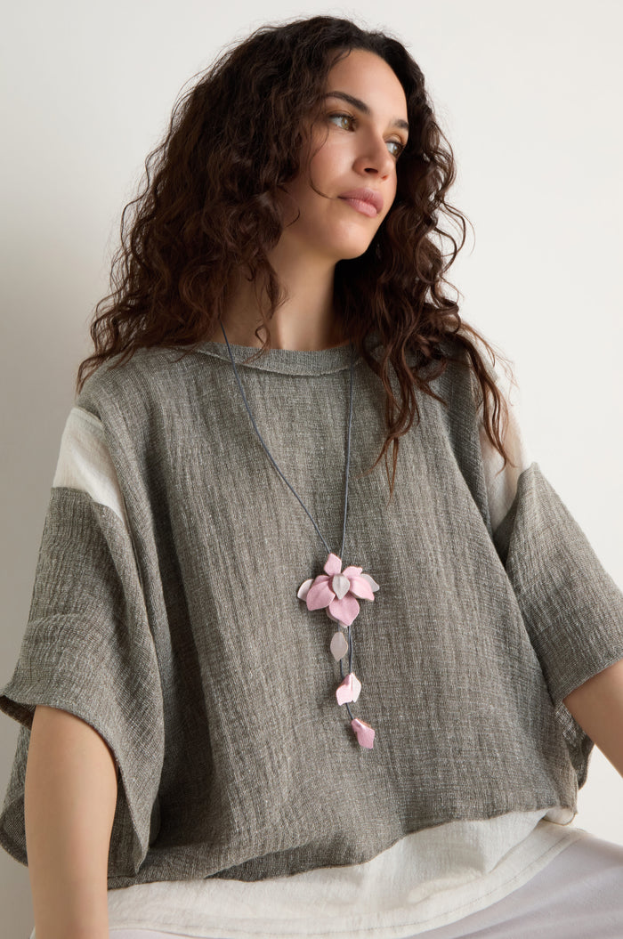 Woman with wavy brown hair wears a loose grey and white top and a long cord necklace featuring the Leather Flower Pendant, which has a handcrafted pink flower, while seated against a plain light background.