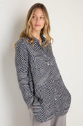 A woman stands against a light background wearing the Linear Prism Shirt, a loose-fitting striped viscose flannel with pockets, paired with light pants, and looks slightly to her left.