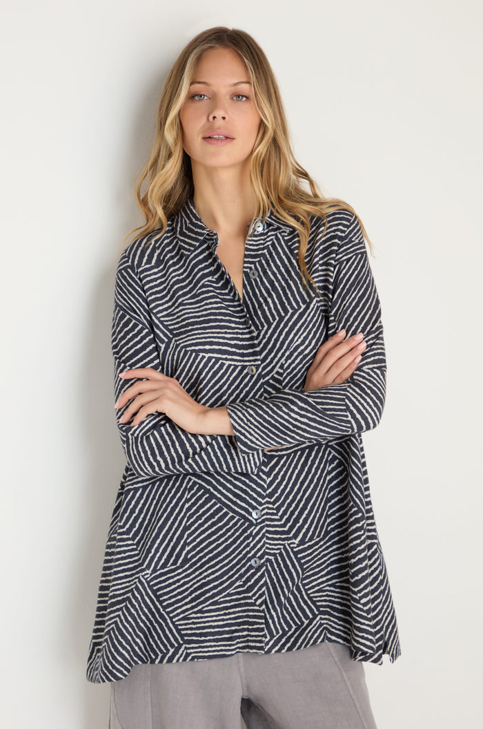 A woman stands against a light background, arms crossed, wearing the Linear Prism Shirt—a long-sleeve top with black and white geometric stripes—paired with light gray pants.