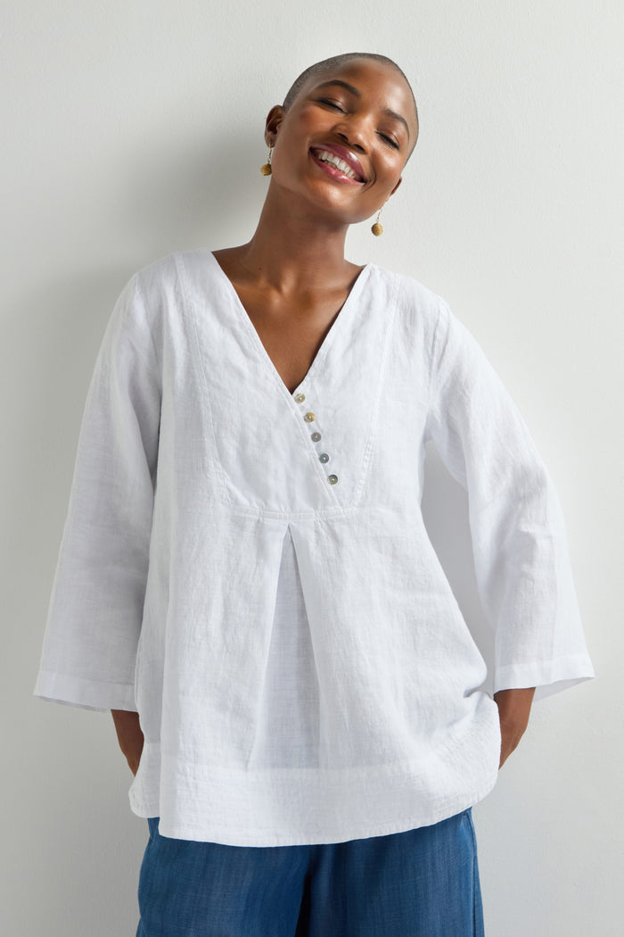 A person wearing a white, long-sleeved blouse styled as a Linen Button Placket Tunic paired with blue pants smiles against a plain background.