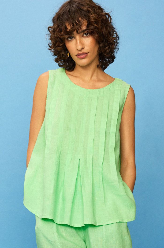 A woman with curly hair wears the Linen Pleat Front Pistachio Vest and matching pleated bottoms, standing against a solid blue background.