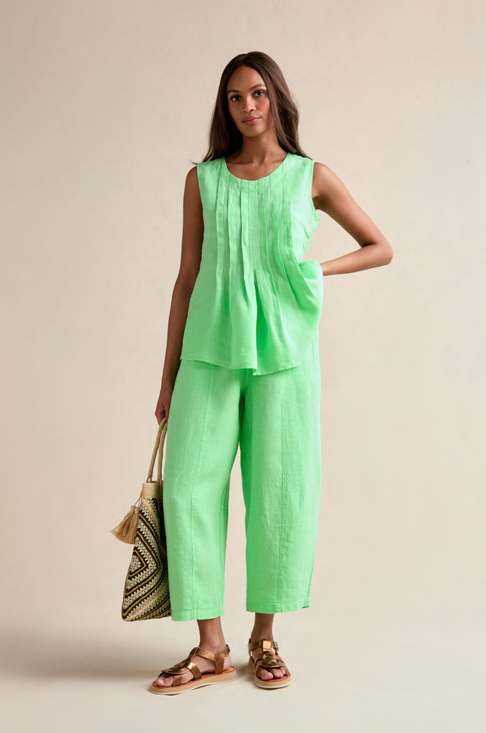 A woman stands against a neutral background wearing the Linen Pleat Front Pistachio Vest with matching wide-leg pants, holding a woven tote bag and wearing brown sandals.