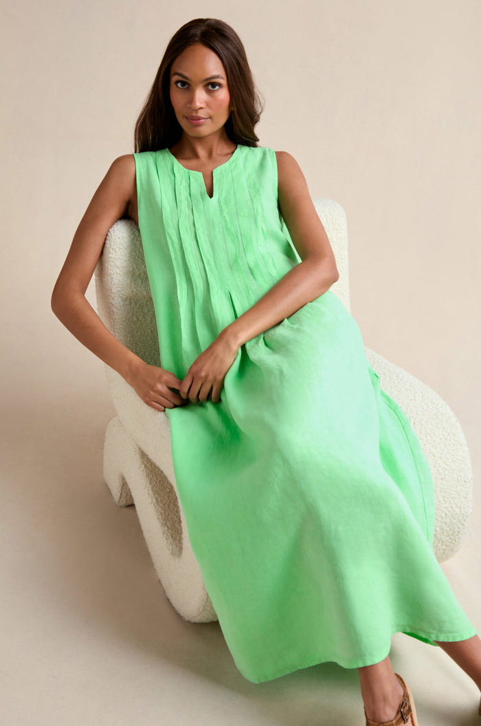 A woman in a light green Linen Pleat Front Sleeveless Dress sits on a white textured chair against a neutral background, showcasing this effortless summer staple.