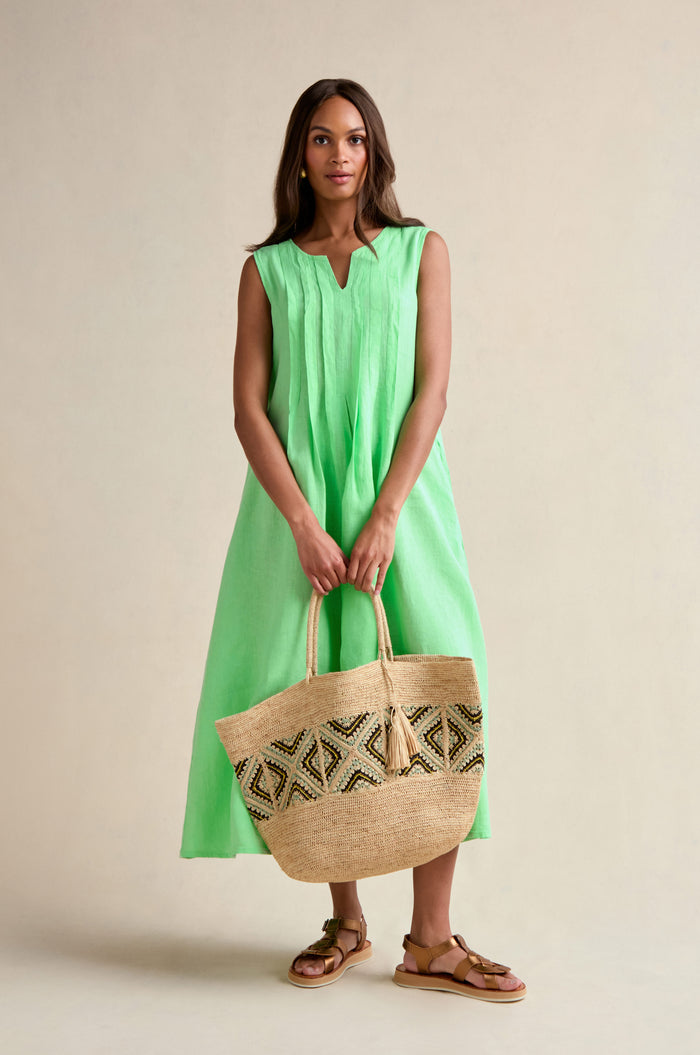 A woman wearing the Linen Pleat Front Sleeveless Dress in mint green holds a large geometric woven tote bag and pairs the look with brown sandals.