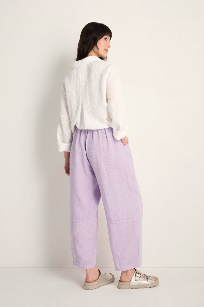 Woman with her back to the camera wears a white textured long-sleeve top, Linen Seam Detail Lilac Bubble Trousers with an elasticated waist, and beige sandals against a plain light background.