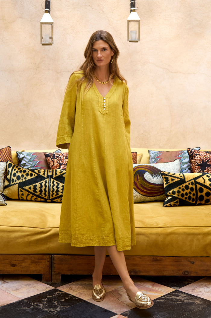 A woman in a Linen Seam Flare Golden Dress and gold shoes stands in front of a patterned sofa with assorted decorative pillows and hanging lanterns above.