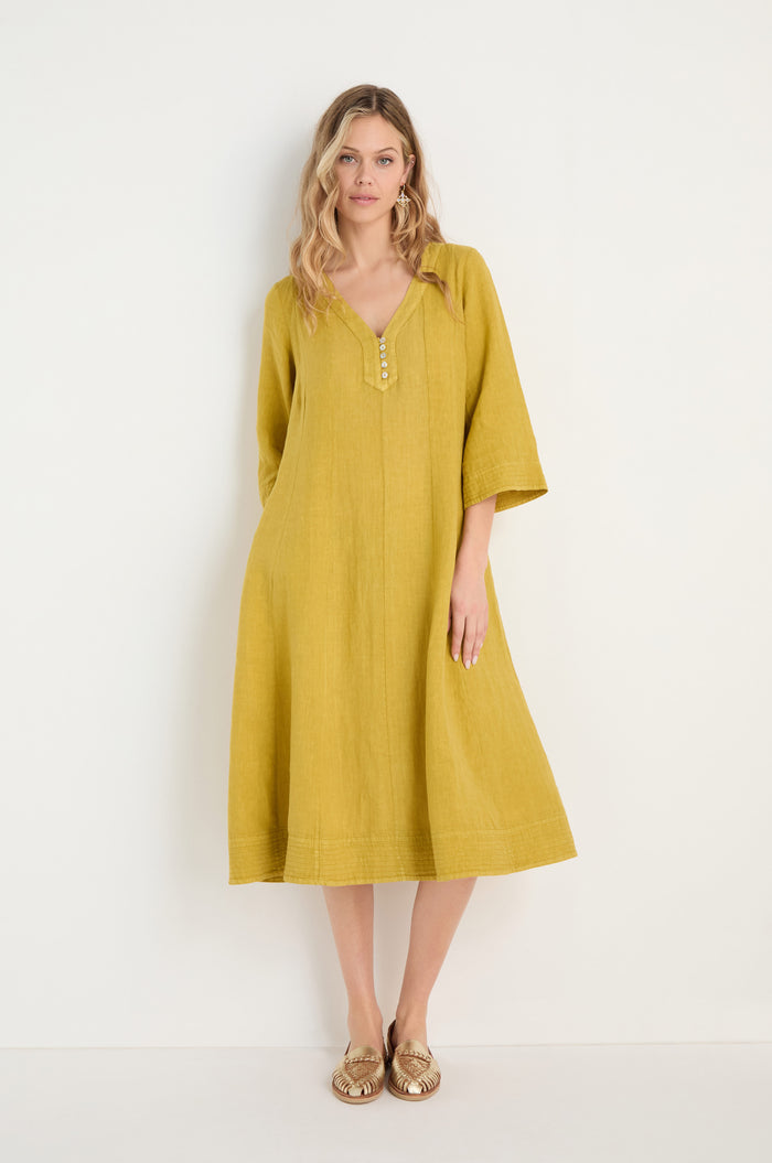 A woman stands against a white wall wearing the Linen Seam Golden Flare Dress—loose-fitting, knee-length, with three-quarter sleeves and subtle panel seams—paired with tan flat shoes.