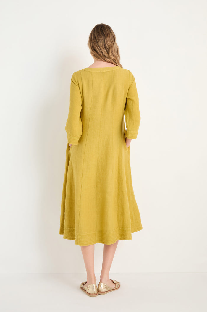 A woman faces away, wearing the Linen Seam Golden Flare Dress—loose-fitting with long sleeves and panel seams—paired with gold sandals against a plain white background.