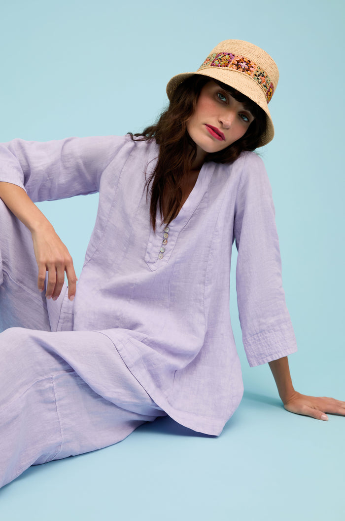 A woman wears the Linen Seam Flare Lilac Tunic and a beige hat with a patterned band while sitting on the floor against a light blue background.