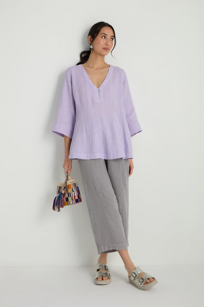 Woman standing against a white wall, wearing the Linen Seam Flare Lilac Tunic with a subtle V-neck, paired with grey cropped pants and sandals, holding a colorful tassel handbag.