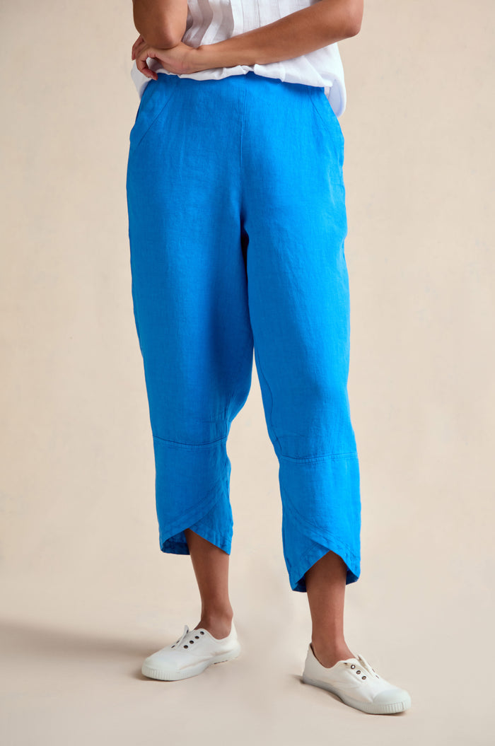 A person wears Linen Tulip Hem Azure Bubble Trouser with a white short-sleeve top and white sneakers, standing with arms crossed against a neutral background.