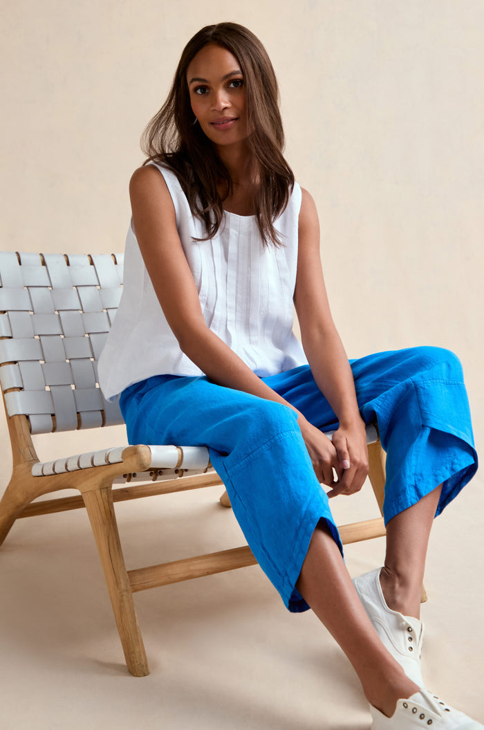 A woman sits on a wooden chair, wearing a sleeveless white top, Linen Tulip Hem Azure Bubble Trouser, and white sneakers against a plain light background.