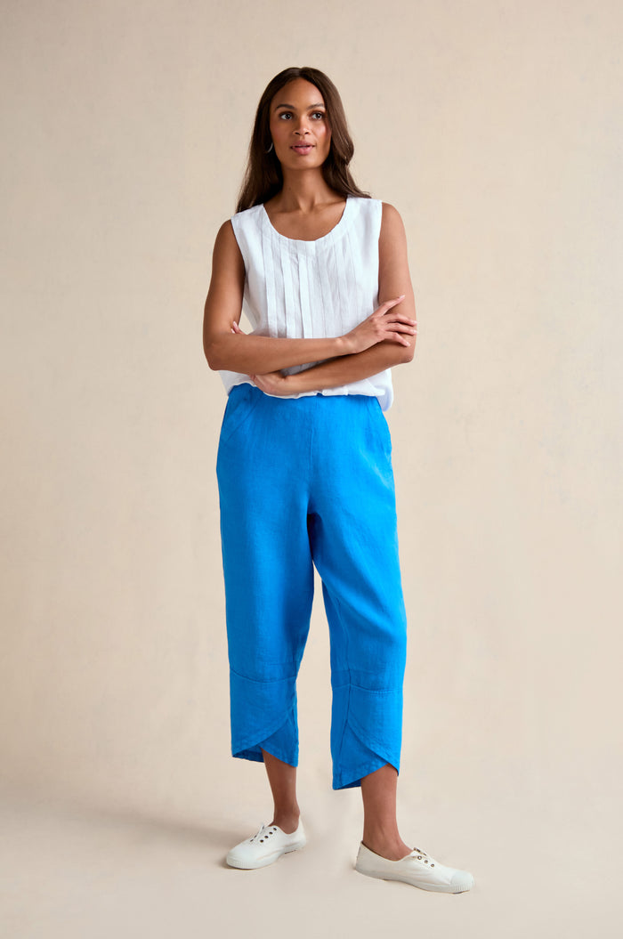 A woman stands with arms crossed, wearing a sleeveless white blouse, Linen Tulip Hem Azure Bubble Trousers cropped at the ankle, and white sneakers against a plain beige background.