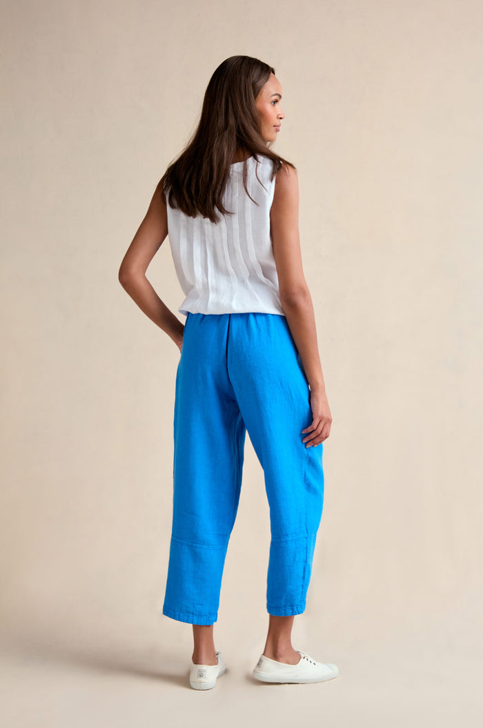 A woman faces away, wearing a sleeveless white top, the Linen Tulip Hem Azure Bubble Trouser in a cropped length, and white shoes against a neutral background.
