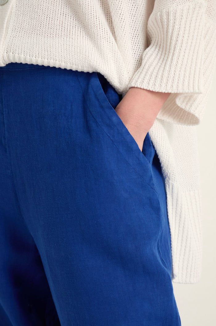 A person wears Linen Tulip Hem Cerulean Bubble Trousers and a cream knit sweater, with one hand in a pants pocket.