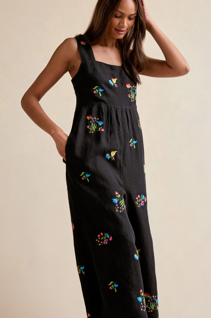 A woman poses against a plain light background, wearing the Vintage Floral Embroidered Jumpsuit featuring sleeveless design and colorful floral embroidery, with one hand touching her head.