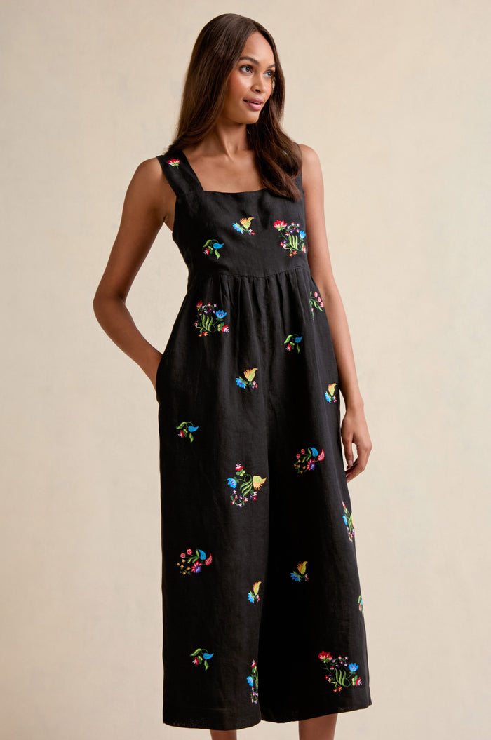 A woman stands against a plain beige background wearing the Vintage Floral Embroidered Jumpsuit, a sleeveless black piece adorned with colorful floral embroidery.