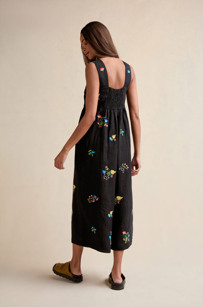A woman stands with her back to the camera on a neutral background, modeling the Vintage Floral Embroidered Jumpsuit—a black, sleeveless piece featuring colorful floral embroidery and a back-smocked design.