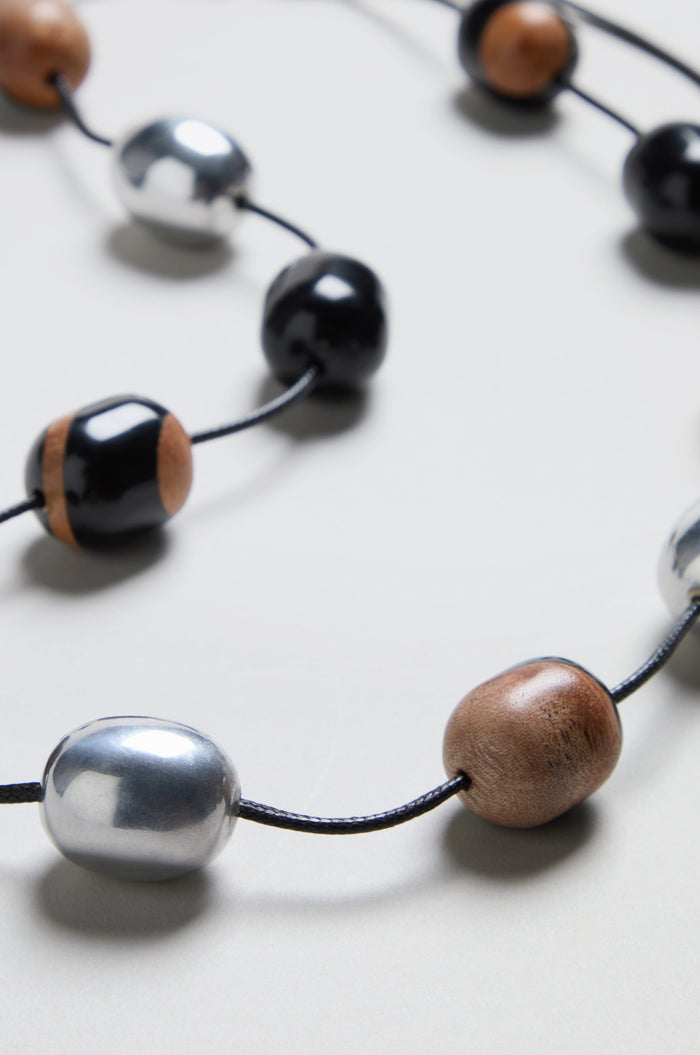 A close-up of the Long Allsorts Necklace, featuring wood and silver elements accented with black, silver, and wood-toned beads elegantly spaced along a black cord resting on a light surface.