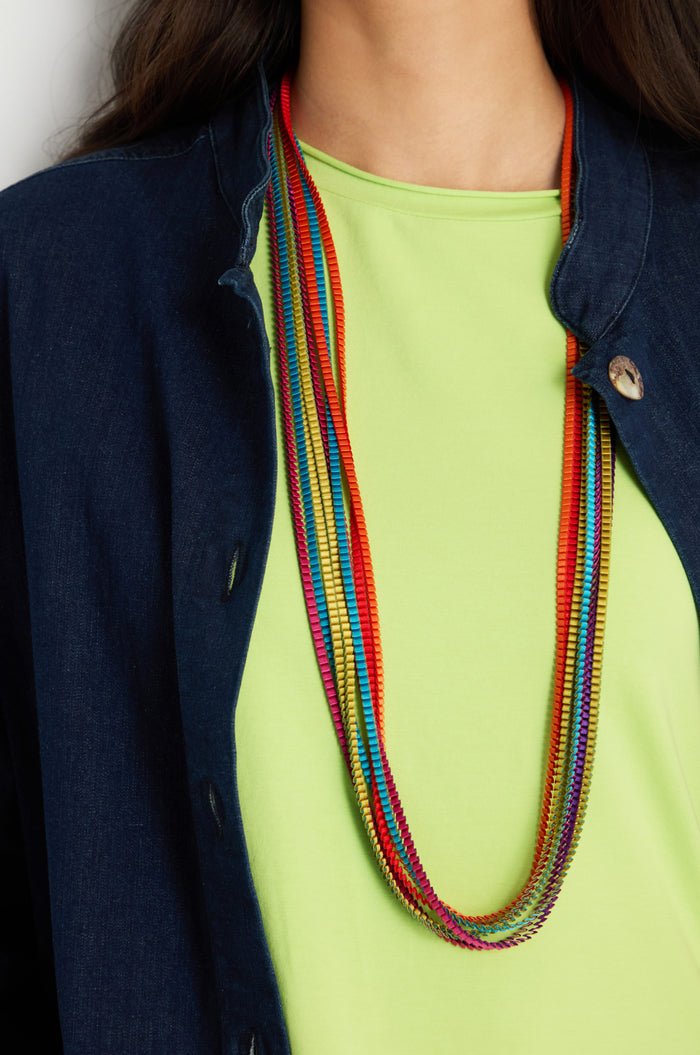 A person wearing a lime green top, a navy blue denim jacket, and the Long Pleated Necklace as a statement accessory.