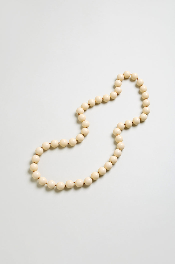 A single strand of round, cream resin beads forms a loose, irregular loop on a light grey background, highlighting the elegant simplicity of the Long Spheres Necklace.