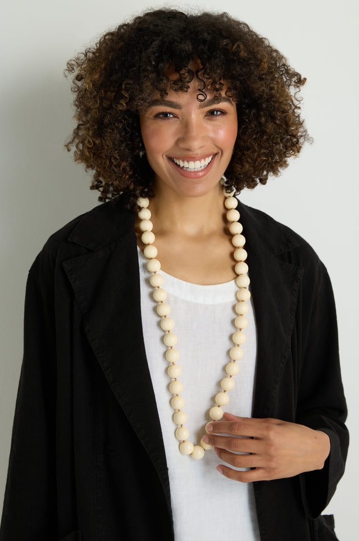 A person with curly hair smiles at the camera, wearing a black jacket over a white shirt and a Long Spheres Necklace featuring large, cream-colored resin sphere beads.