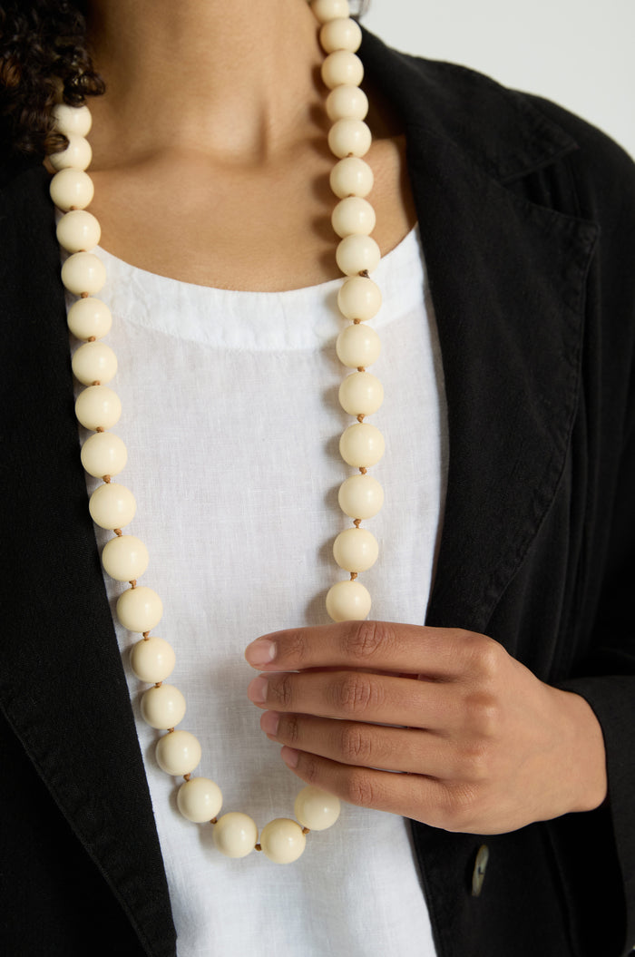 Wearing the Long Spheres Necklace with cream resin sphere beads over a white top and black blazer, this person is holding the necklace in one hand.