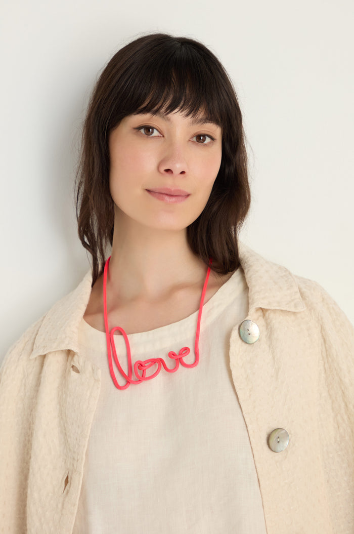 A woman with medium-length dark hair wears a cream textured jacket and the Love Necklace, a handmade accessory featuring "love" in red script.