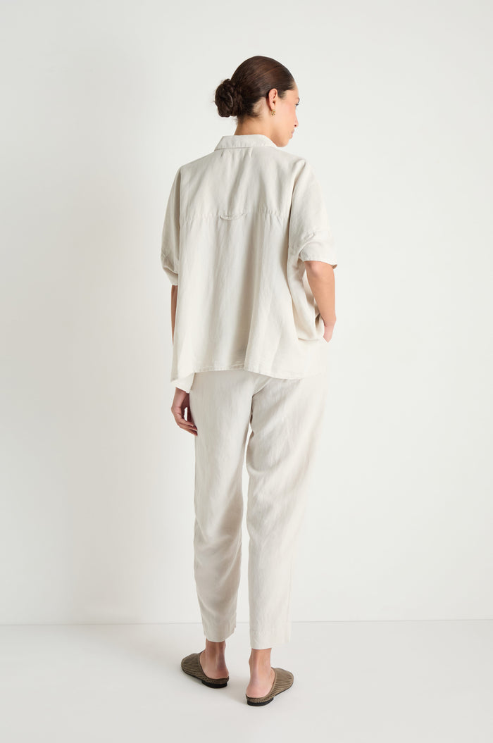 A person stands facing away, wearing the Lyocell Linen Boxy Shirt in a relaxed fit and light color, paired with matching pants. Their hair is tied back and they wear dark slip-on shoes.
