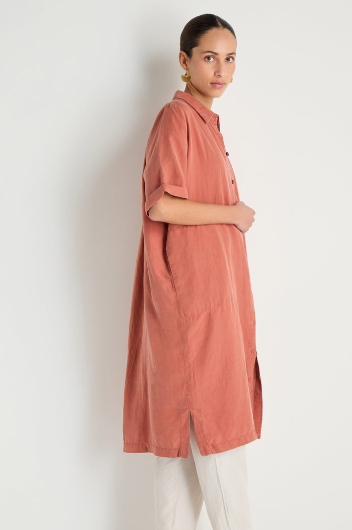 A person stands sideways wearing the Lyocell Linen Shirt Dress in a relaxed fit with short sleeves and white pants against a plain light background.