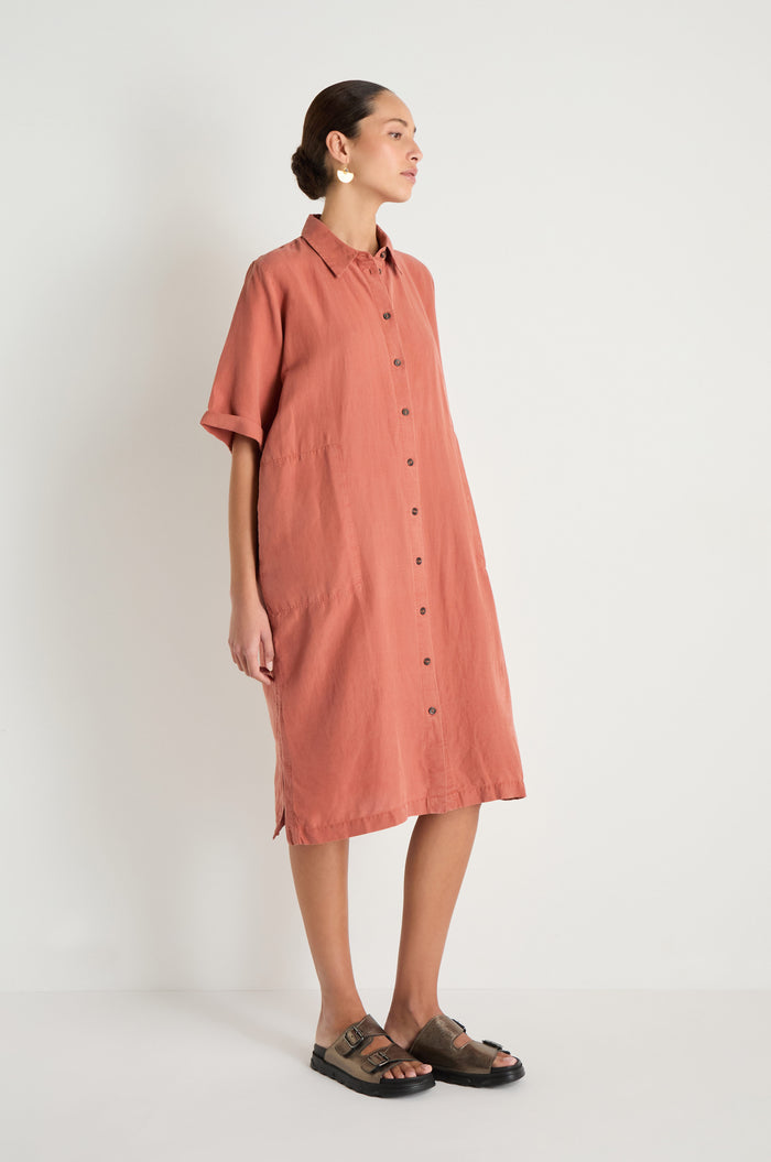 Person stands against a white wall, wearing the Lyocell Linen Shirt Dress—a loose, button-up, short-sleeve rust dress—paired with black sandals and circular earrings.