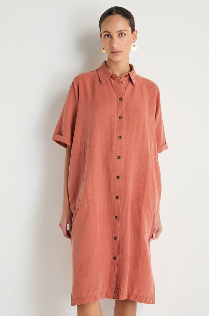 A woman stands against a plain background wearing the Lyocell Linen Shirt Dress in rust, featuring a relaxed fit, button-through front, short sleeves, and side pockets.