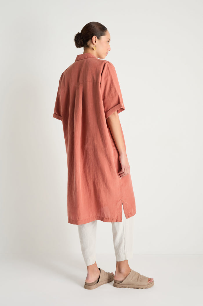 A woman faces away against a plain background, wearing the Lyocell Linen Shirt Dress in rust, styled as a relaxed-fit, short-sleeved shirt dress over light pants and beige slide sandals, highlighting the back of the outfit.