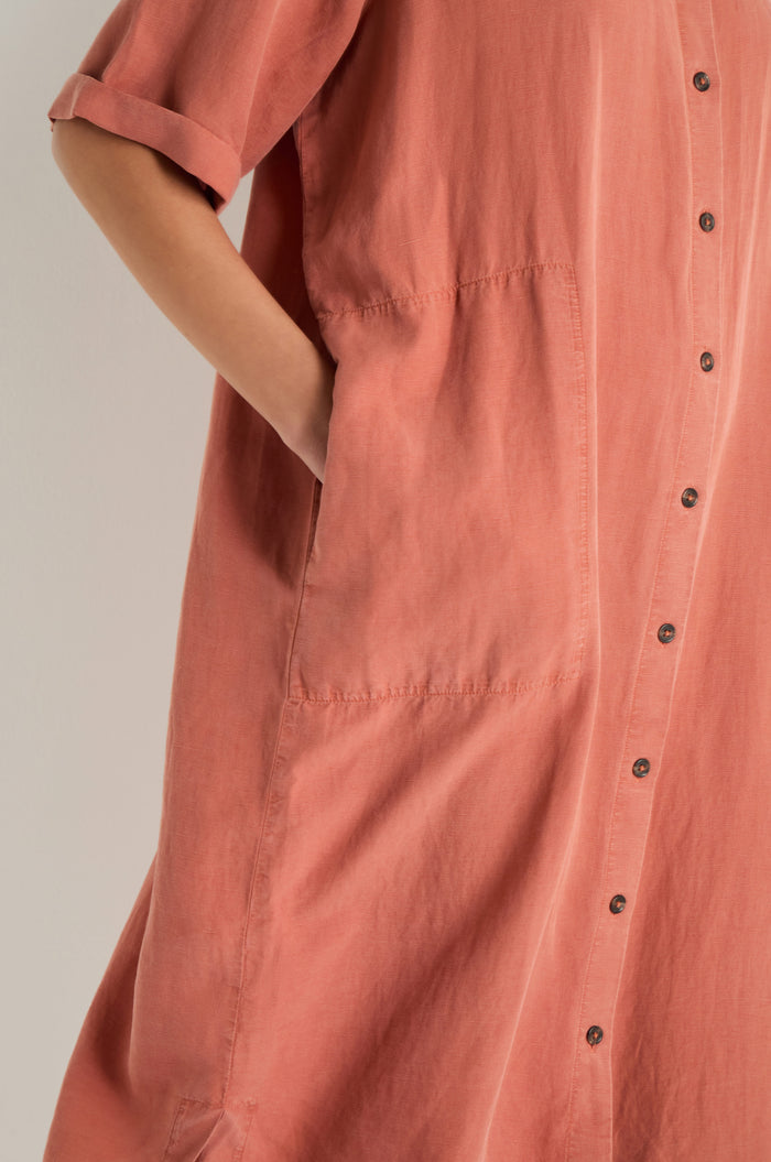 A person wears the Lyocell Linen Shirt Dress in muted pink, featuring a relaxed fit, button-through front, short sleeves, and large side pockets.