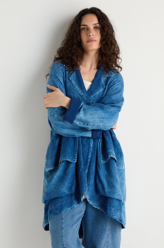 A person with long curly hair stands against a white wall, arms crossed, wearing the Lyocell Utility Jacket in blue over a white top and blue denim pants.